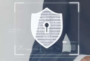 En arrière plan on voit le corps d'un homme en costume qui utilise une tablette tactile. Au premier plan, on aperçoit le symbole du bouclier qui fait ici référence à la cyberassurance.